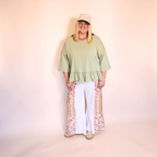 A woman modeling a apple green ruffled short-sleeve top, white wide-leg pants with colorful geometric patterned side panels, and a beige baseball cap against a solid pale pink background.