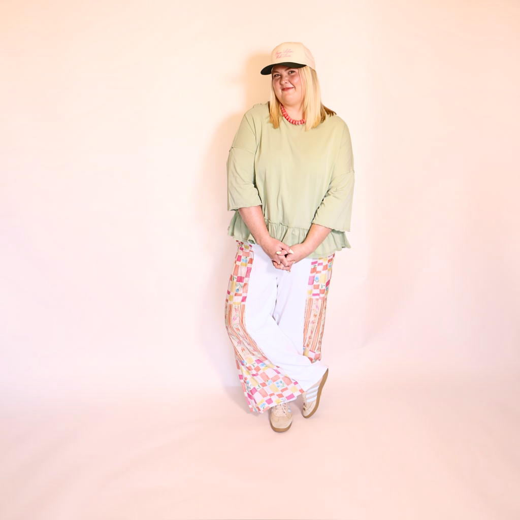 A woman modeling a light apple green oversized t-shirt, white pants with colorful geometric patterned side panels, a beige baseball cap, and beige sneakers, standing against a solid pale pink studio backdrop.