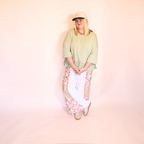 A woman modeling a light apple green oversized t-shirt, white pants with colorful geometric patterned side panels, a beige baseball cap, and beige sneakers, standing against a solid pale pink studio backdrop.