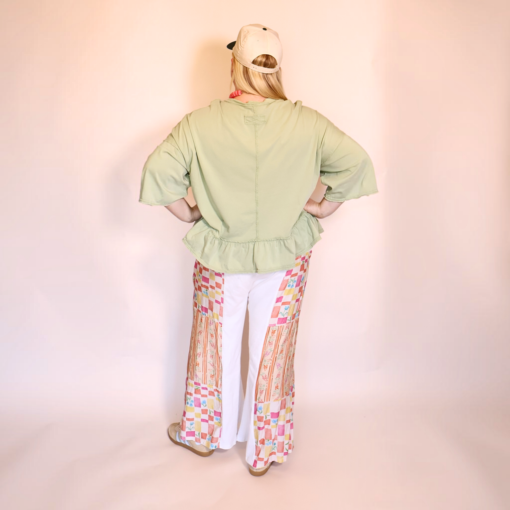 A person shown from the back wearing a light apple green peplum blouse, white pants with colorful geometric patterned side panels, and a beige baseball cap against a pale pink studio backdrop.
