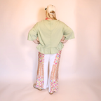 A person shown from the back wearing a light apple green peplum blouse, white pants with colorful geometric patterned side panels, and a beige baseball cap against a pale pink studio backdrop.