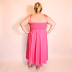 A woman viewed from the back wearing a bright pink midi dress with thin shoulder ties and a smocked bodice, standing against a plain light pink studio backdrop.