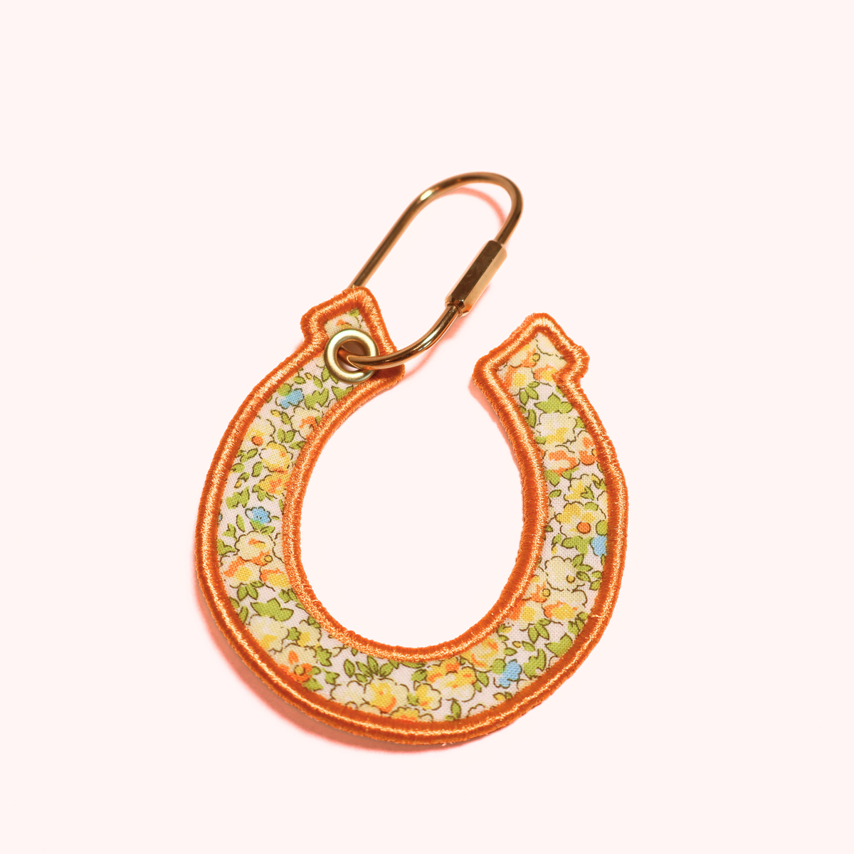 A horseshoe-shaped keychain or bag charm with a floral fabric pattern in yellow, green, and blue, finished with an orange embroidered border and a gold-tone metal carabiner clip, shown on a solid pale pink background.