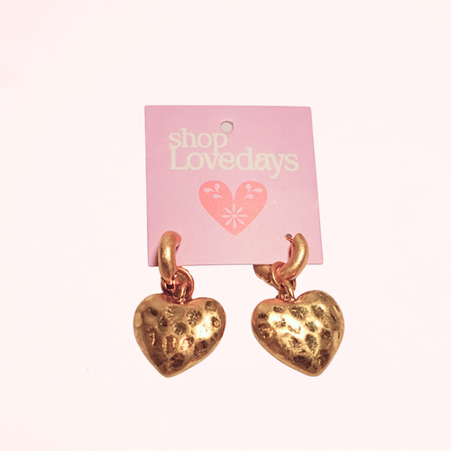 A pair of textured gold heart-shaped dangle earrings hanging from a pink display card printed with 'shop Lovedays'.