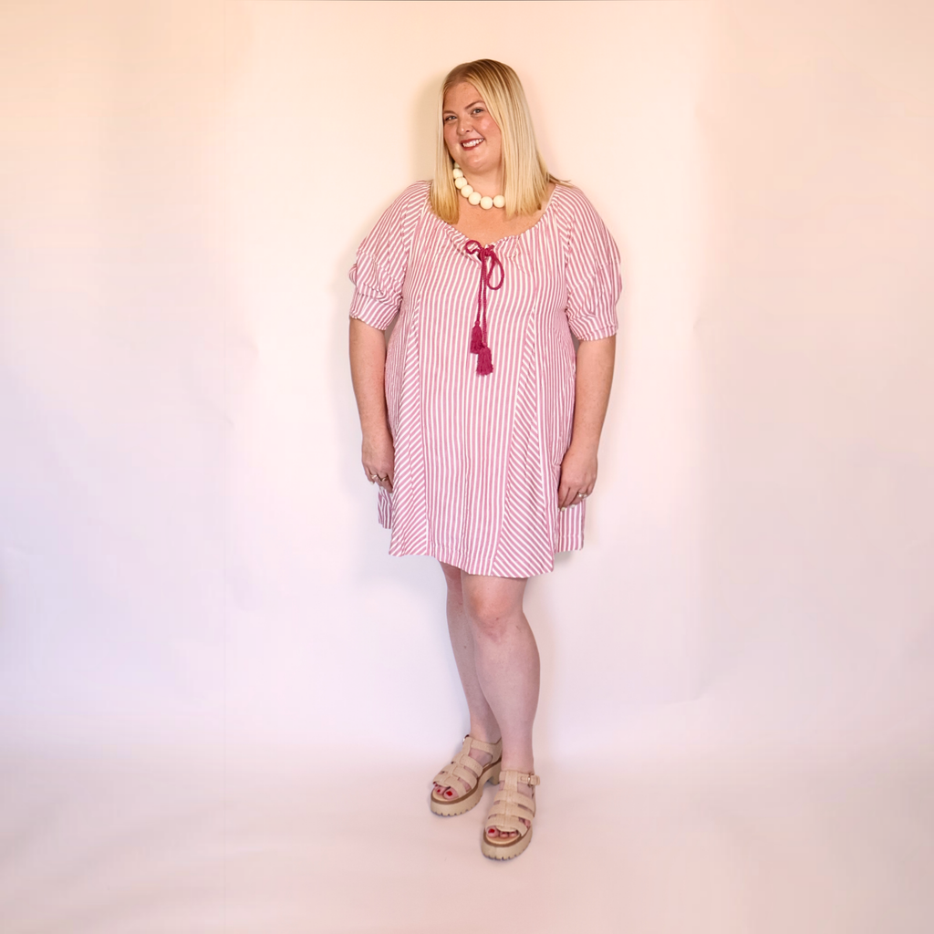 A woman modeling a pink and white vertically striped short-sleeve tunic dress with a dark red tassel tie at the neckline, styled with a white beaded necklace and beige platform sandals against a plain light background.