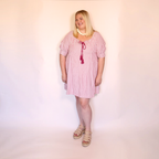 A woman modeling a pink and white vertically striped short-sleeve tunic dress with a dark red tassel tie at the neckline, styled with a white beaded necklace and beige platform sandals against a plain light background.