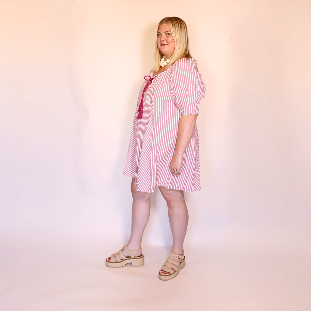 A woman modeling a pink and white vertically striped short-sleeve dress with a pink bow detail at the neckline, paired with beige platform sandals, standing against a plain light pink studio backdrop.