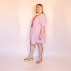 A woman modeling a pink and white vertically striped short-sleeve dress with a pink bow detail at the neckline, paired with beige platform sandals, standing against a plain light pink studio backdrop.