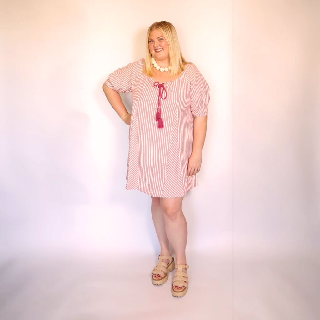 A woman modeling a pink and white vertically striped short-sleeve tunic dress with a magenta tassel tie at the neckline, styled with a white beaded necklace and beige platform sandals against a plain white studio background.