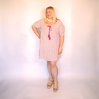 A woman modeling a pink and white vertically striped short-sleeve tunic dress with a magenta tassel tie at the neckline, styled with a white beaded necklace and beige platform sandals against a plain white studio background.