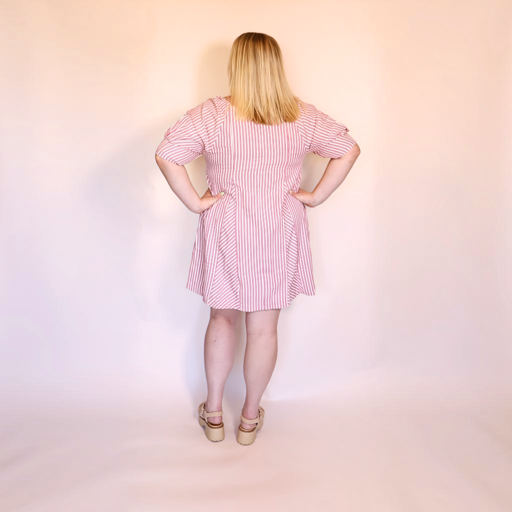 A woman stands with her back to the camera wearing a pink and white vertically striped short-sleeve dress with a smocked waist, paired with beige platform sandals, against a plain light pink studio backdrop.