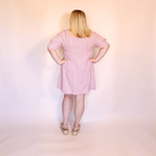 A woman stands with her back to the camera wearing a pink and white vertically striped short-sleeve dress with a smocked waist, paired with beige platform sandals, against a plain light pink studio backdrop.
