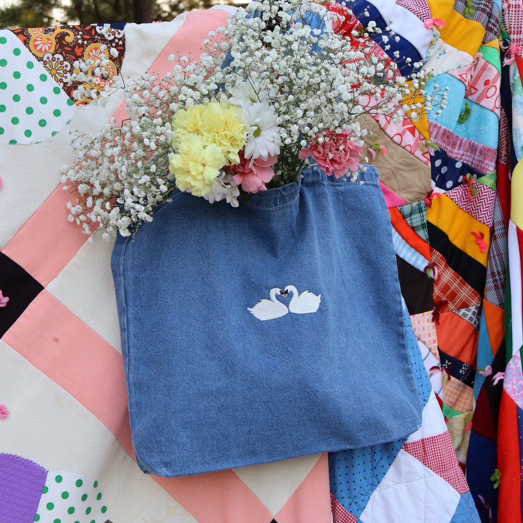 A blue fabric tote bag with a white swan design, filled with baby's breath and flowers, resting on a colorful patchwork quilt.
