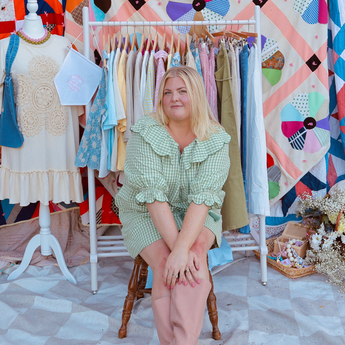 A woman sits on a wooden chair in front of a colorful quilt display. She is wearing a light green ruffled dress