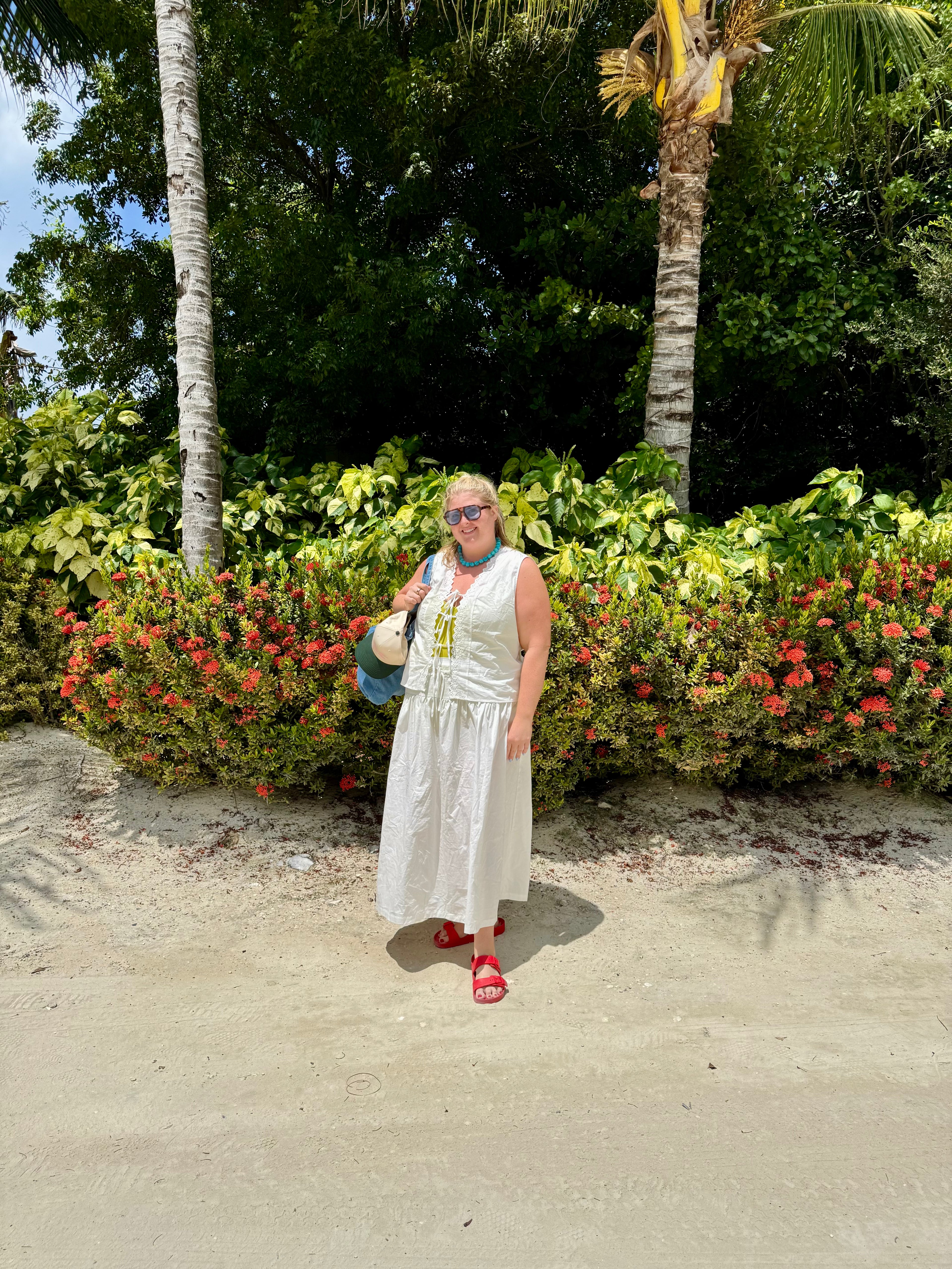 A woman wearing a white sleeveless top and skirt and red slide sandals stands on a sandy path surrounded by tropical green foliage and red flowers.