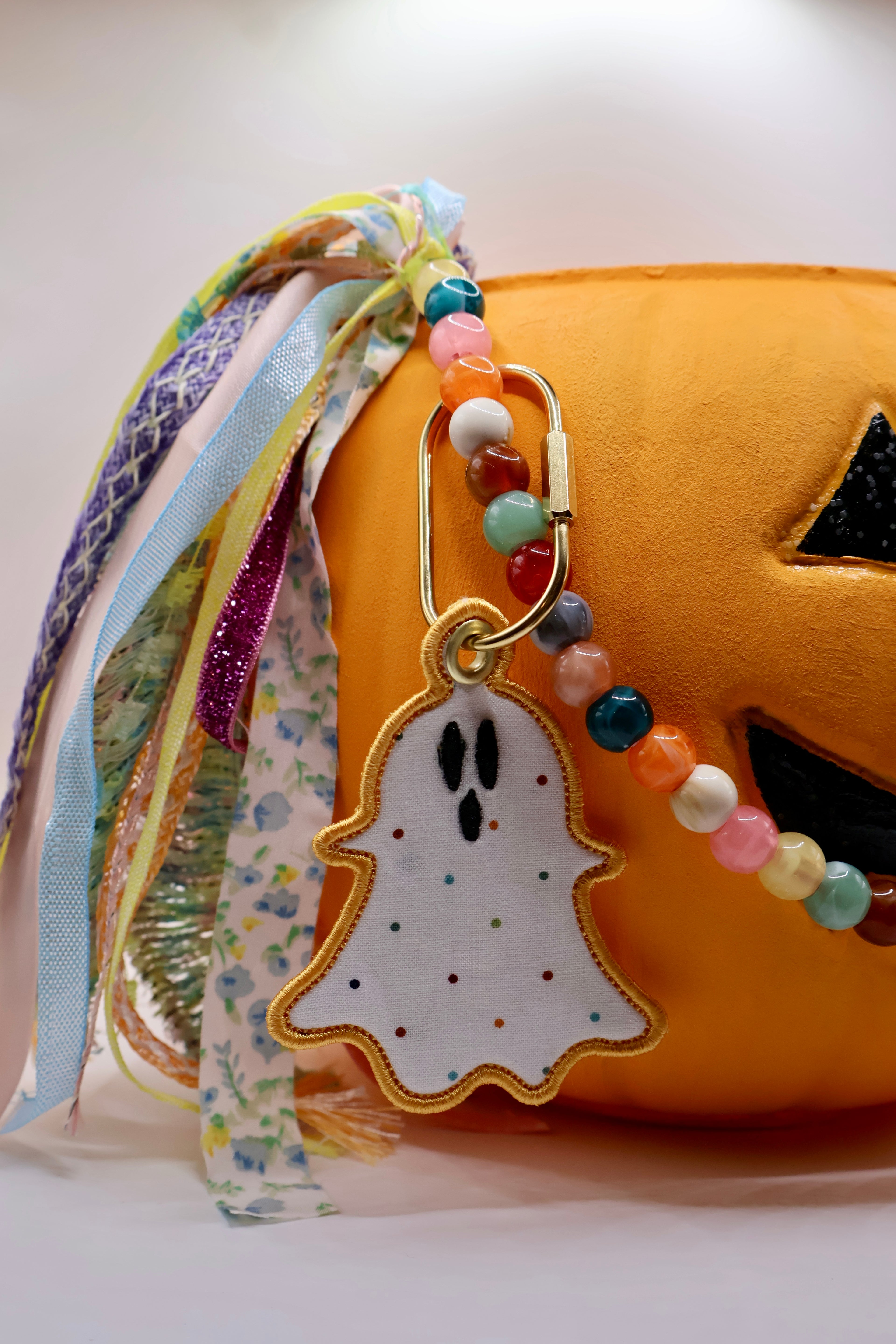 A Halloween-themed keychain featuring a white ghost-shaped charm with embroidered eyes and mouth, attached to a gold clasp. The clasp is connected to a beaded strap with colorful round beads and draped over a plush orange pumpkin, with multiple patterned fabric ribbons hanging on the left side.